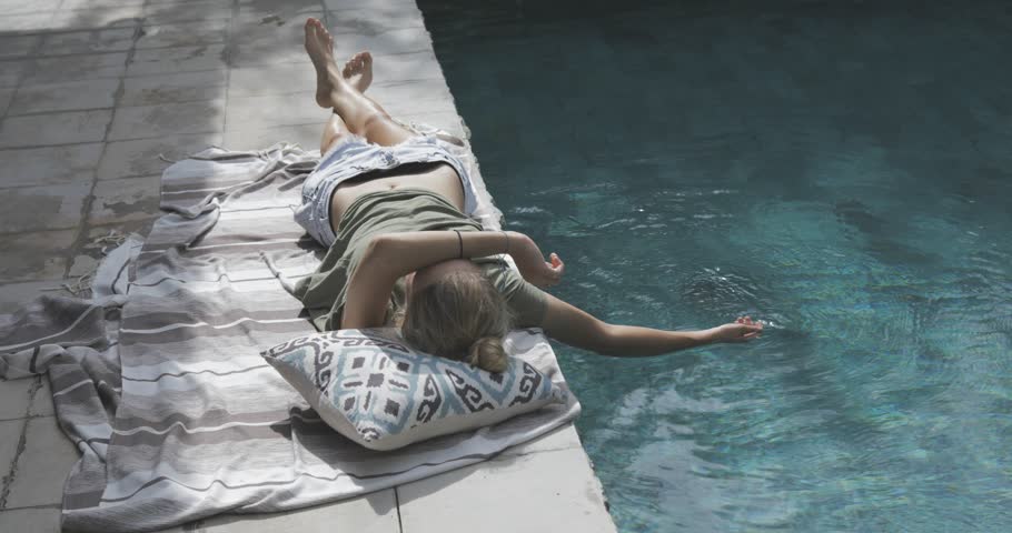 fit tanned good looking nice girl or young woman resting near swimming pool in the backyard, holding and refreshing her hand in water, tree shadow , sunny warm weather