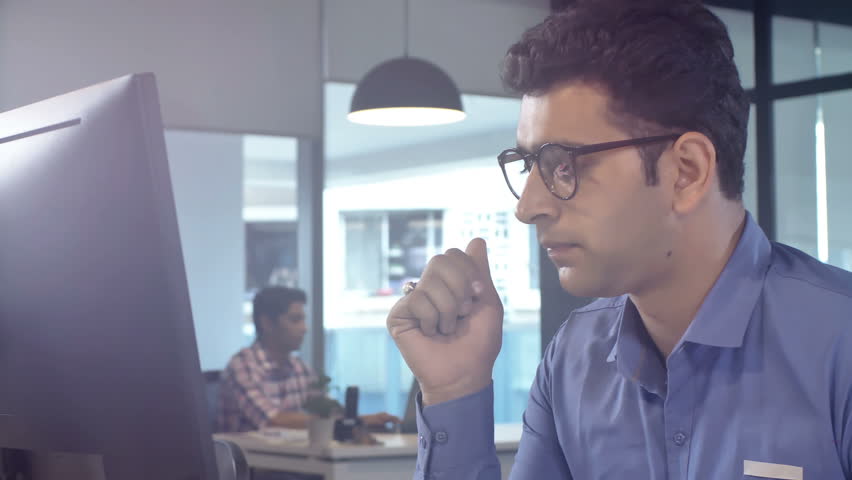 Moving shot of a Stressed out, overburdened and tired out young male staff member sitting in front of a computer screen removes his eye glasses in modern corporate office Royalty-Free Stock Footage #1008814163