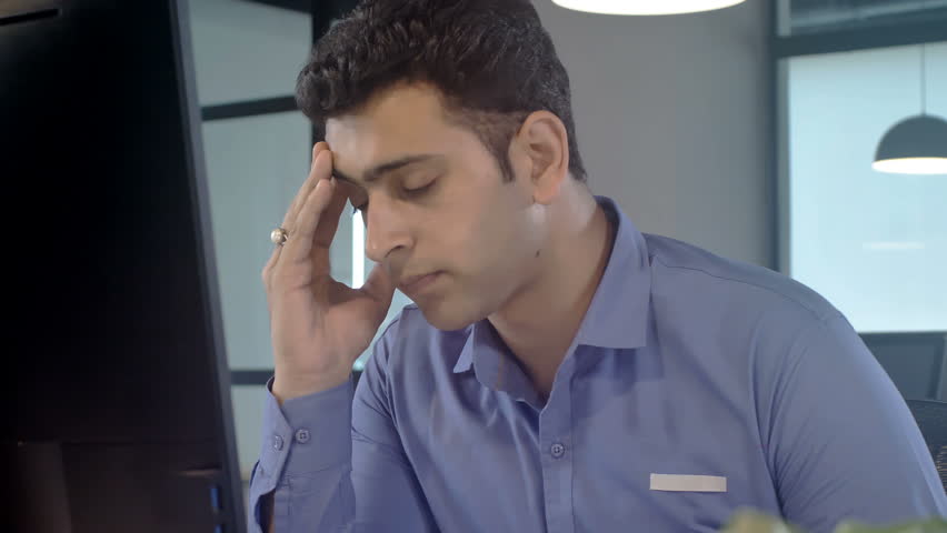 Moving shot of a Stressed out, overburdened and tired out young male staff member sitting in front of a computer screen removes his eye glasses in modern corporate office | Shutterstock HD Video #1008814163
