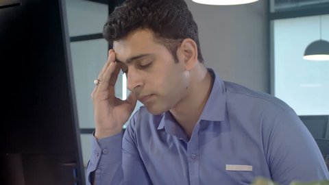 Moving shot of a Stressed out, overburdened and tired out young male staff member sitting in front of a computer screen removes his eye glasses in modern corporate office