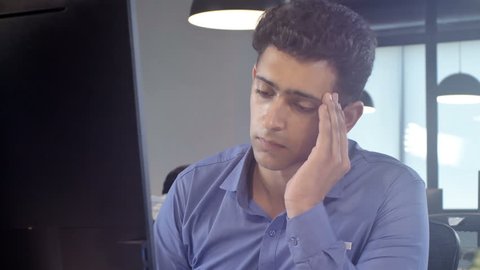 Track shot of a overworked, tired and exhausted young male office employee removes his eye glasses sitting in front of the computer screen in a modern corporate office 