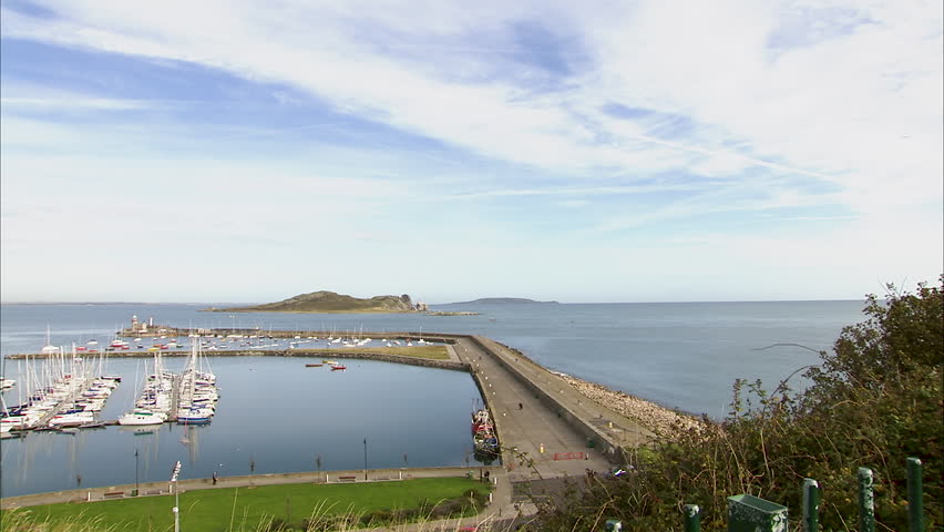 Wide panning shot of a man made bay in Ireland, with many yachts docked, green hills, houses and a blue cloudy sky.