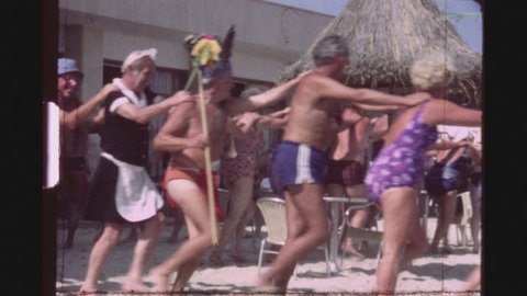 SPAIN, MALLORCA, JULY 1983. A Group Of Female And Male Caucasian Tourists Dancing Happily In A Conga Line Circle In Bathing Suits At A Beach Bar.