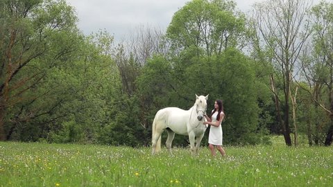 Attractive young girl is walking with white unicorn in the woods. She is wearing light white dress.