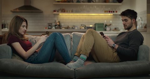 Man and woman ignore each other on the sofa while using their smartphones for social media.