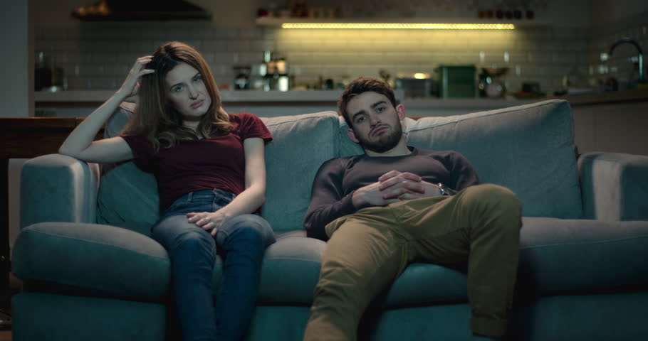 Extremely bored and frustrated couple sitting on a sofa at home struggle to watch a terrible movie.. | Shutterstock HD Video #1008855278