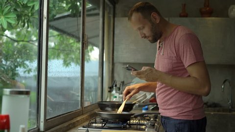 Young man using smartphone while cooking meal in kitchen 
