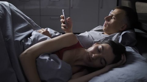 Young married couple lying in bed at night. Nervous man using cellphone texting with lover, while his wife is sleeping. When woman wakes up, man hastily hides his smartphone and embraces his wife.