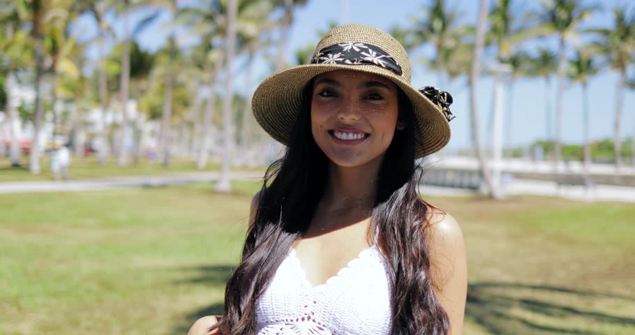 Pretty young model wearing trendy straw hat and white dress touching hair and posing at camera on tropical resort. 