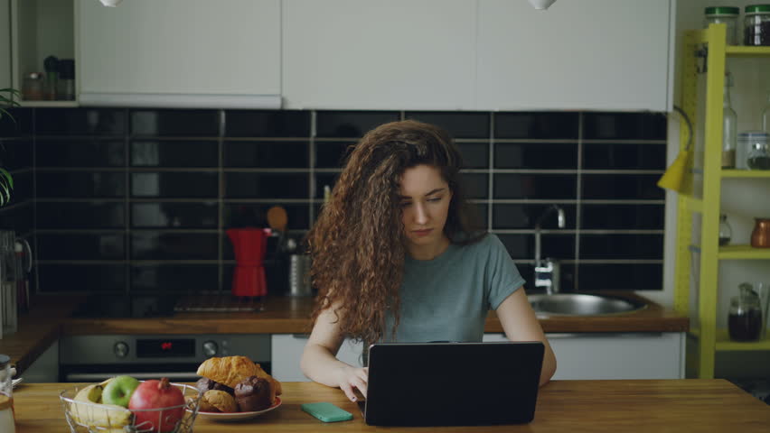 Young positive caucasian curly womn sitting ar table in kitchen working on laptop, her husband comes and she shows him something in laptop, they are smiling and discussing it