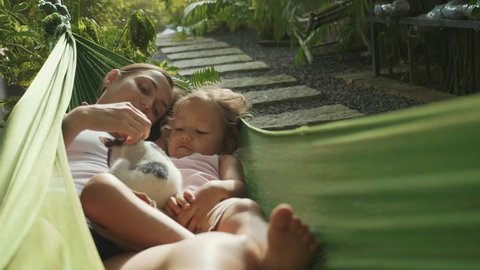 Happy mother and daughter relaxing together in a hammock with cat at garden in sunny summer day, slow motion. Family playing with kitty in hammock.