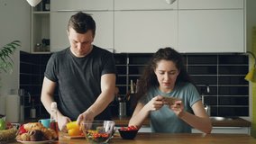 Attractive couple in the kitchen at home. Curly girl playing video game on smartphone while her boylfriend cooking