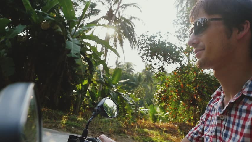Young smiling man traveler driving motorbike on tropical sunny road. slow motion. 1920x1080