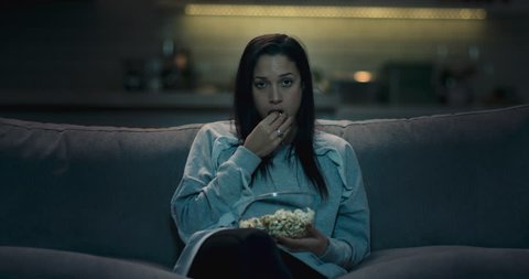 Sad, bored and depressed woman sitting on a sofa at home watching a bad movie eating popcorn