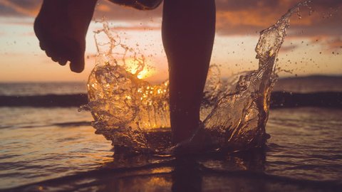 SLOW MOTION, CLOSE UP, COPY SPACE: Woman jogger splashes water by running into the refreshing ocean at sunrise. Picturesque golden sunbeams slowly disappear behind horizon as woman runs into the sea.