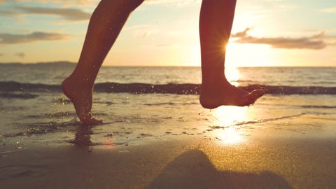 SLOW MOTION, CLOSE UP: Unrecognizable young woman runs barefoot along beautiful sandy beach at sunset. Playful female traveler jogs on warm sand near the vast blue ocean on breathtaking summer evening