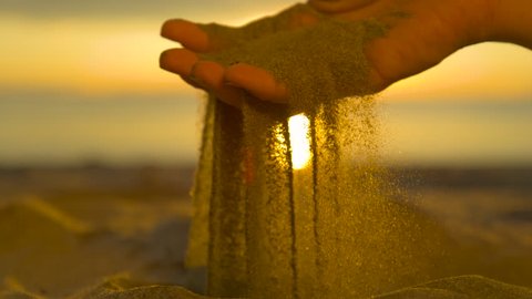 SLOW MOTION CLOSE UP, REVERSED: Small particles of dry sand fall between female fingers and back on shore. Beautiful summer sunset illuminates sandy beach and woman sifting dry sand with her hands.