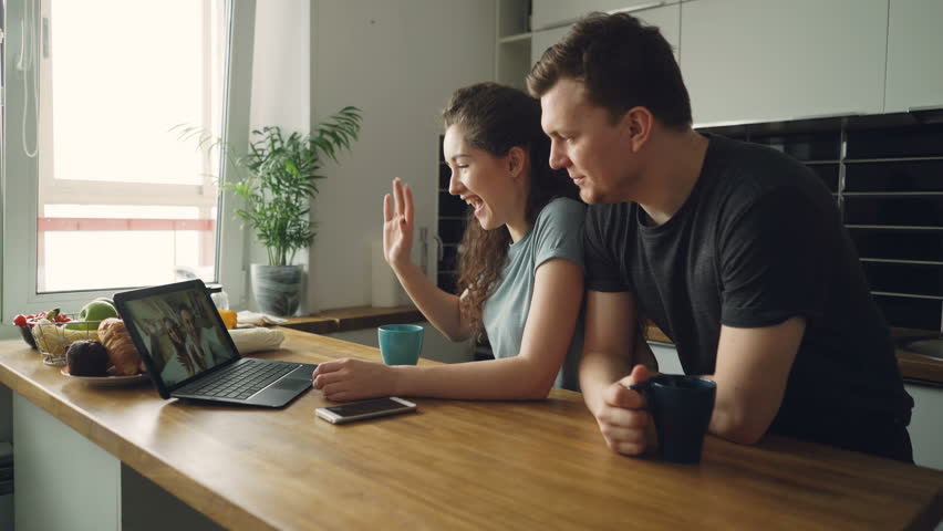 Young cheerful caucasian couple sitting at table in front of laptop video chatting with their happy positive tanned friends from abroad, they are finishing conversation waving hands | Shutterstock HD Video #1008926021