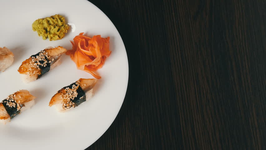 Large white plate on which lies four huge sushi with eels rewound with a piece of nori next to pink ginger and green wasabi