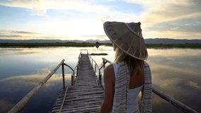 Young woman wearing Asian conique hat contemplating the beautiful sunset from a lake bridge, beautiful reflection on water surface. People travel Asia relaxation concept, shot in 4K resolution video.