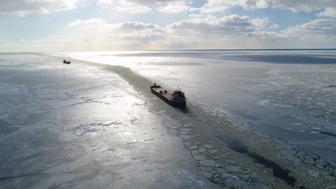 Cargo ship sailing on frozen sea in extreme winter conditions aerial shot. Sailing in narrow fairway channel made by icebreaker vessel. Water transportation during cold winter season in north.
