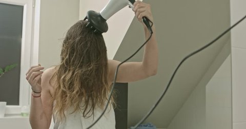 Casual woman with long brown hair drying hair with electric blowdryer.