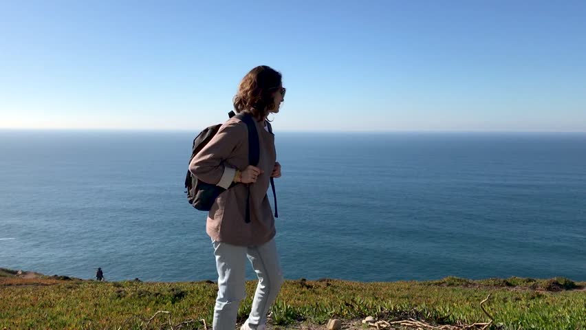 A young woman walks away from the camera along a path on a hill on a background of the blue ocean. A girl with a backpack turns around, looks at the camera and smiles.