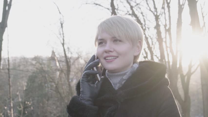 young nice girl speaking by mobile phone in winter park. slow motion. sun background