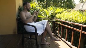Young man in towel chatting on smartphone sitting on terrace 
