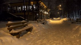 Snowfall in the city park on a winter evening. Benches, building and road covered with heavy snow. HD video