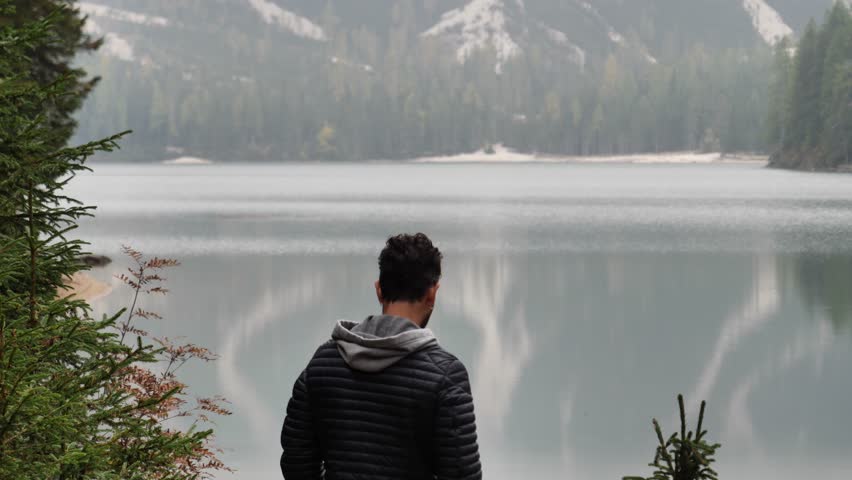 Handsome yung man standing by mountain lake, on background of woods looking away. Braies lake or Pragser Wildsee in Trentino region, Italy