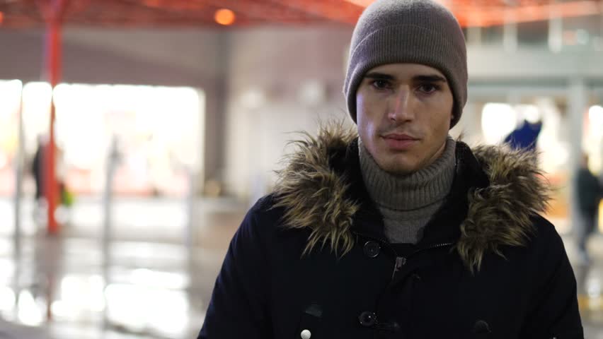 Handsome trendy happy young man standing outdoor at night in city setting in front of big shop window, looking at camera with a smile