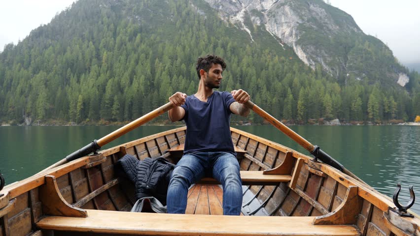 Young casual man sitting in wooden boat and rowing while looking away on background of lake and mountains. Braies lake or Pragser Wildsee in Trentino region, Italy