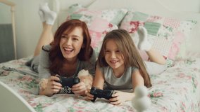 Beautiful happy mother with little daughter laughing and have fun while playing computer console games on TV lying on bed at home in the morning in cozy bedroom
