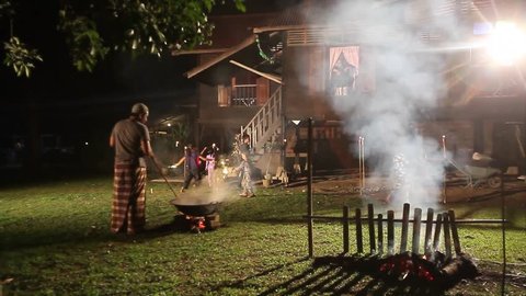 Men making Malaysian traditional food 'lemang' over firewood outdoor kitchen during Eid Mubarak with kids playing firework around vintage village house.Eid ul-Fitr night scene.