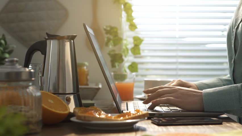 Woman having breakfast in the kitchen, she is connecting with a laptop and receiving messages on her smartphone