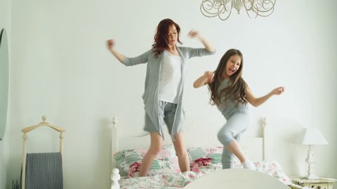 Happy family of cute daughter and young mother jumping and dancing on bed while have fun during morning on holidays at home