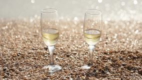 Two glasses of champagne stand on the sand with small shells by the sea, in the background waves wash the shore