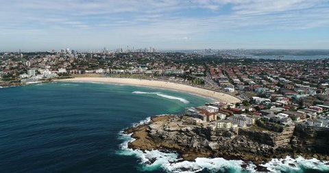 Rocky north bondi head approaching from open sea towards sandy stripe of beach with distant Sydney city CBD landmarks.
