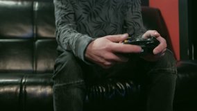Portrait of attractive brunet hipster with beard, holding joy stick and playing videogames on tv at home, sits on the cozy leather black couch, successful, he is winning