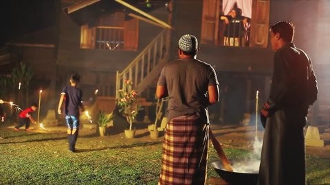 Men cooking 'dodol' (sweet toffee confection) during festival such as Eid ul-Fitr and Eid ul-Adha with kids playing fire cracker in the background at night.