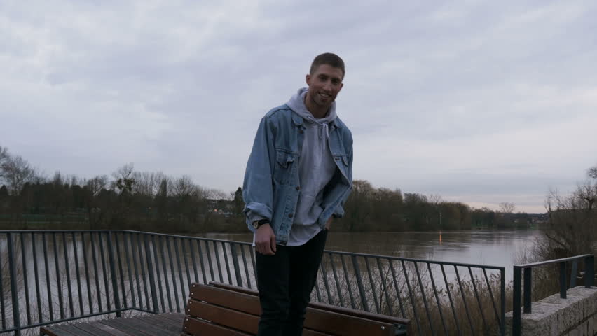 Happy young man standing on a bench in natural parkland with the Main River flowing in the background. Portrait. Three quarter length.