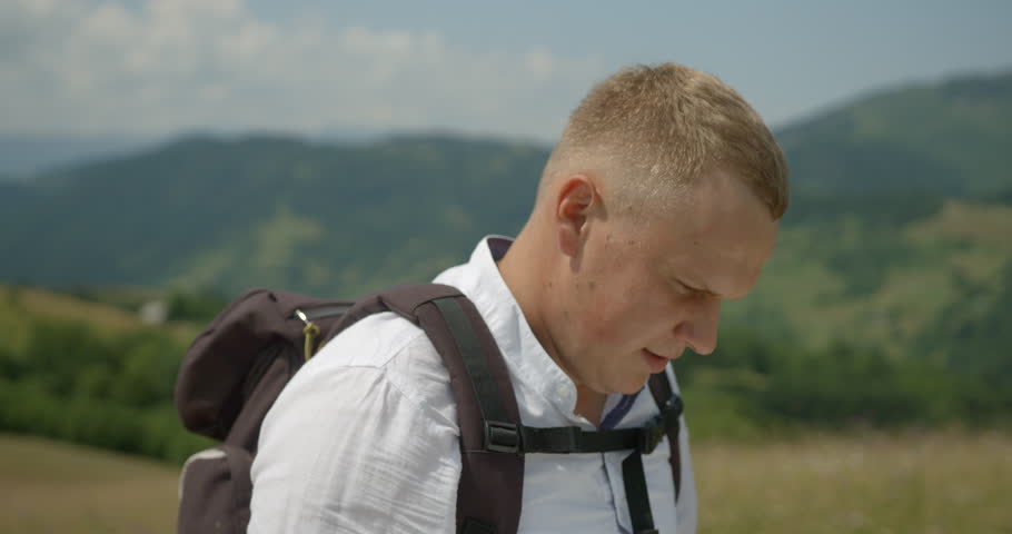 Close-up side portrait of the handsome happy blond man tourist with backpack checking the road with the paper map during hiking in mountains. 4k footage.
