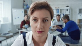 Medium shot of woman with short haircut greeting someone during video conversation and showing her colleagues, who are working in the background