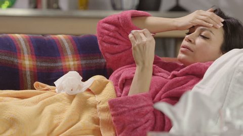 Woman home sick with cold, flu and fever looking at thermometer