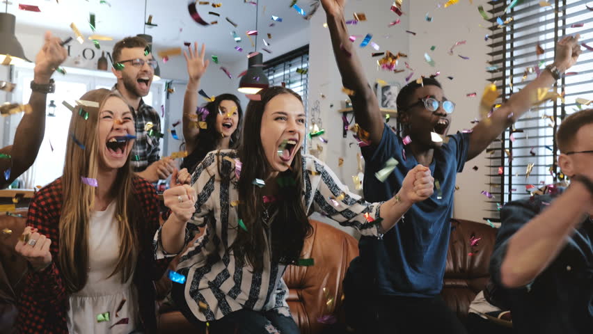 Emotion. Multi-ethnic fans celebrate winning. Confetti 4K slow motion. Passionate supporters shout watching game on TV.