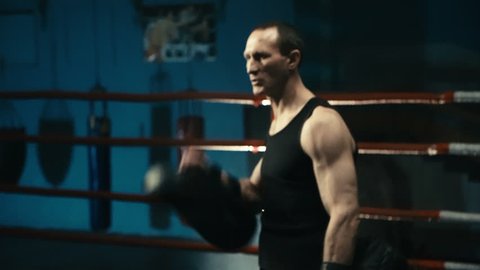 Handheld shot of shirtless muscular boxer with tattooed body working out on ring in gym attacking coach with punching bags.