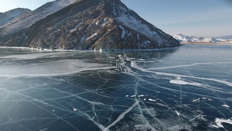 Scenic blue clear transparent ice Lake Baikal. Smooth surface deep cracks. Tourists people walk travel. Cinematic  High mountains snow cliff. Best Russia Buryatia. Winter day blue sky. Aerial Above
