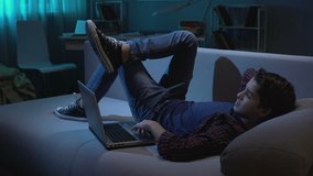 Teenage boy lying on sofa, looking around and watching adult videos on laptop