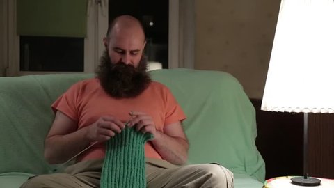 A charismatic man with a beard knits a green scarf while sitting in a room on the couch. Bearded man trying to knit